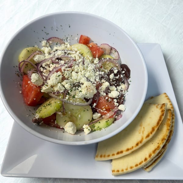 A white bowl of Greek salad with feta, tomatoes, cucumbers, and red onions, served with pita bread.