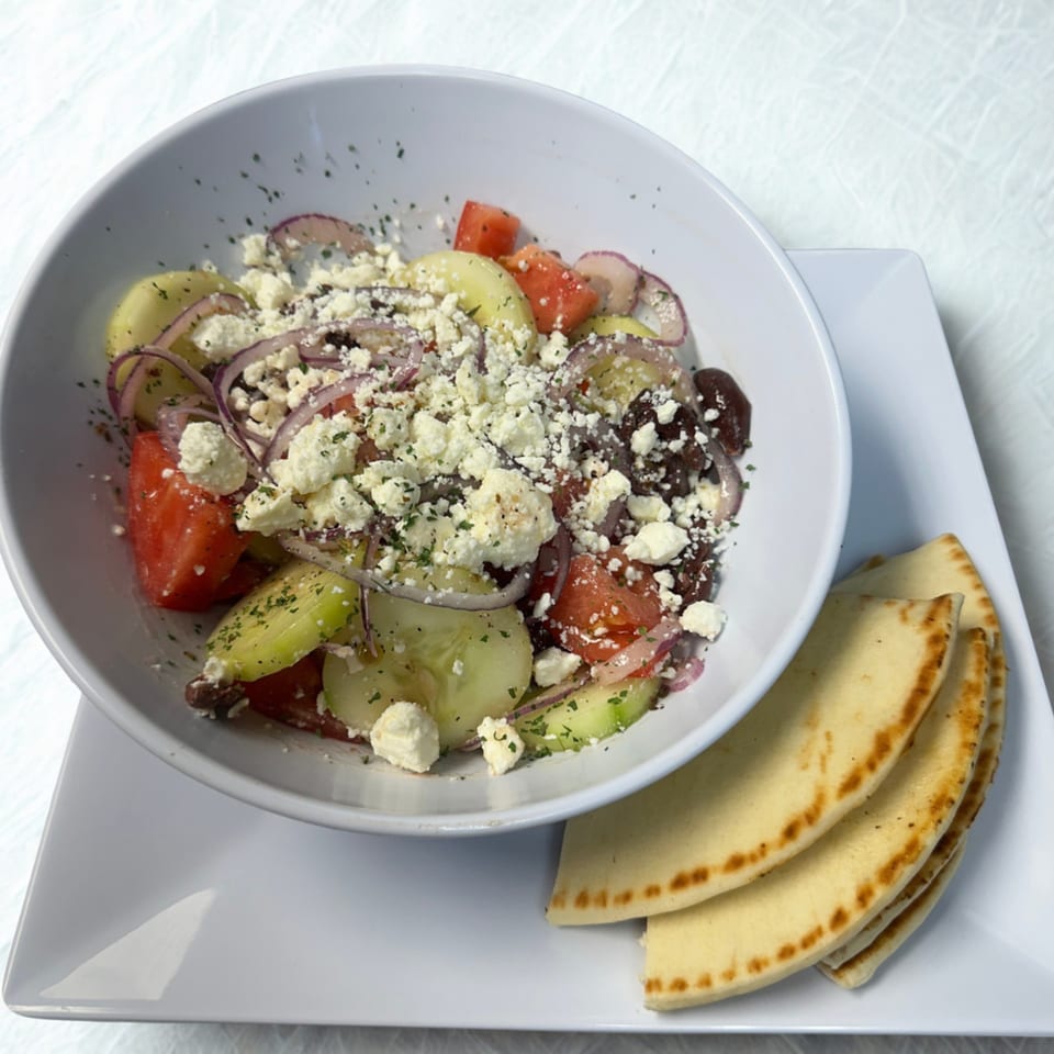 A white bowl of Greek salad with feta, tomatoes, cucumbers, and red onions, served with pita bread.