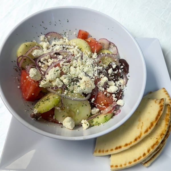 A white bowl of Greek salad with feta, tomatoes, cucumbers, red onions, and olives, with pita bread.
