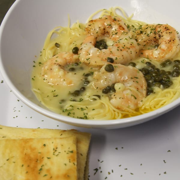 Shrimp scampi with capers and parsley over spaghetti in a white bowl, with a side of garlic bread.