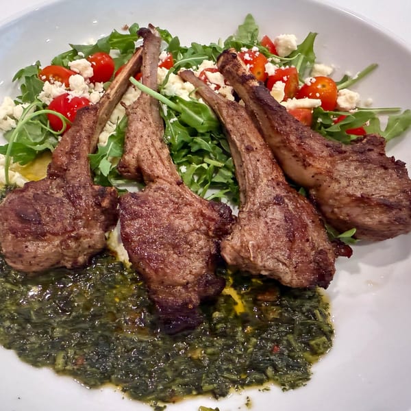 Four grilled lamb chops with a side of green salad, cherry tomatoes, feta cheese, and a green sauce.