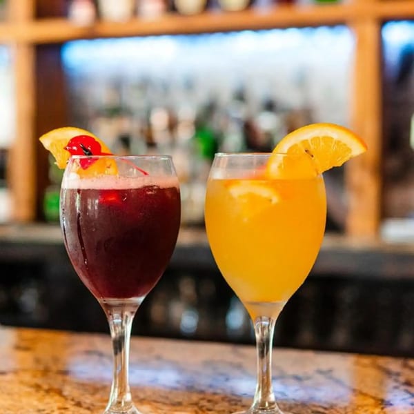 Two cocktails, one red and one orange, garnished with fruit on a bar top at Spiro's Taverna.