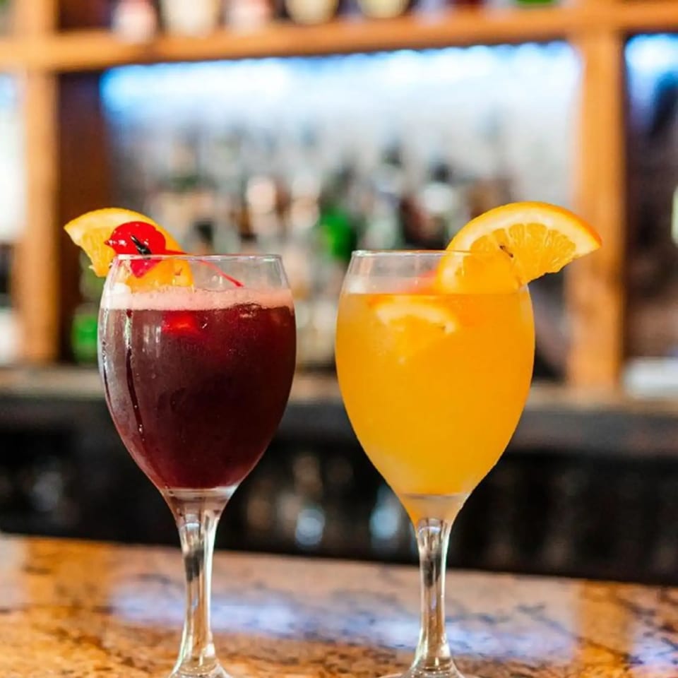 Two cocktails, one red and one orange, garnished with fruit on a bar top at Spiro's Taverna.