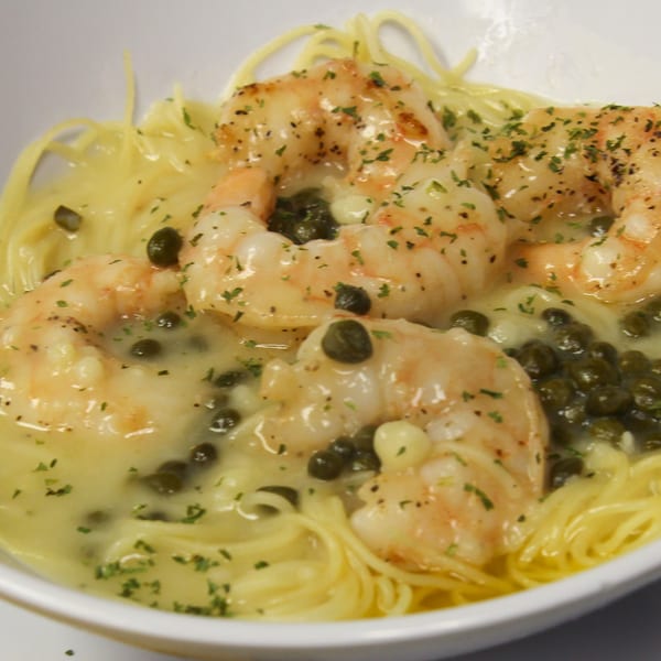 Shrimp scampi with capers and parsley over spaghetti in a white bowl.