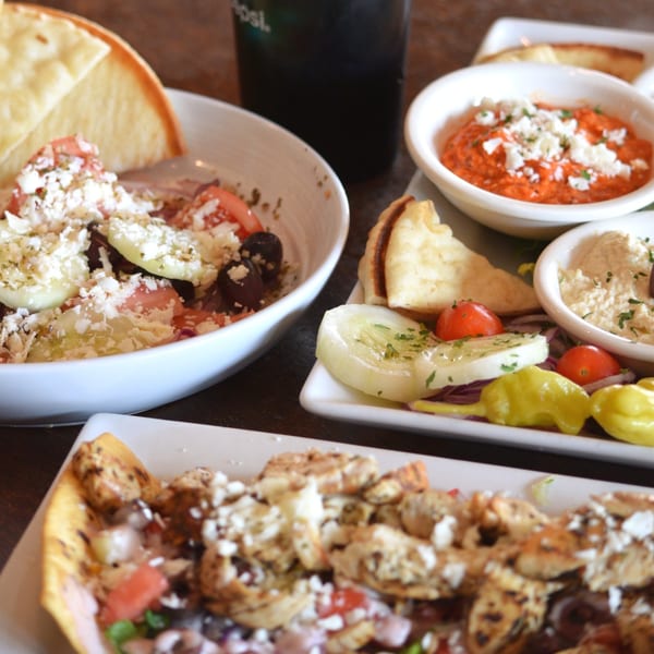 A table laden with Greek dishes, including salad, hummus, pita bread, and a chicken entree.